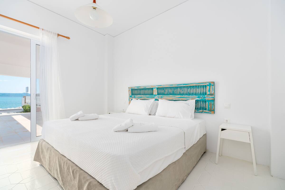 Un dormitorio con cama, cortinas blancas, una mesita de noche blanca y vista al mar.