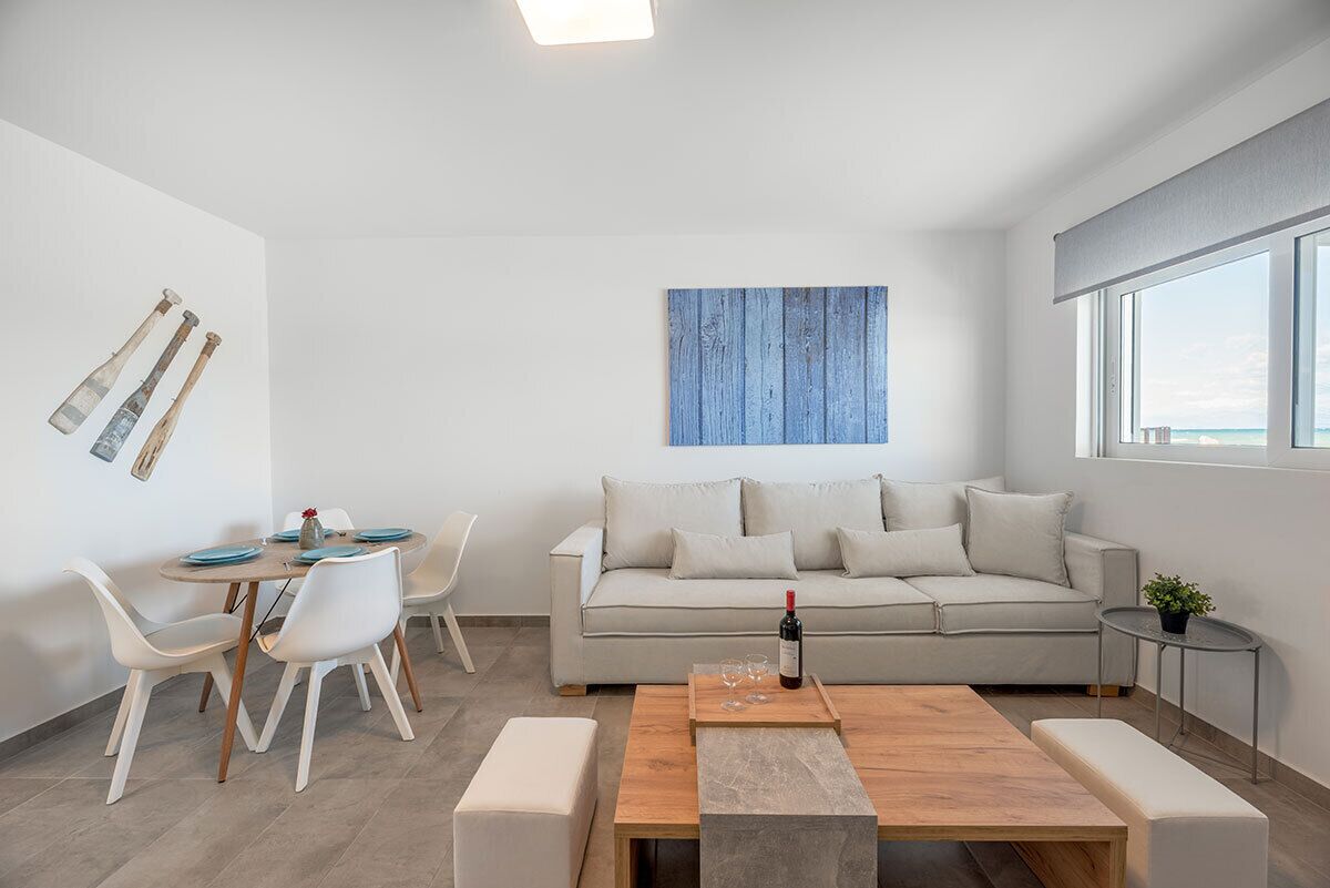 A modern living room with a sofa, a dining table, and chairs. There is a painting on the wall and a window with a view.