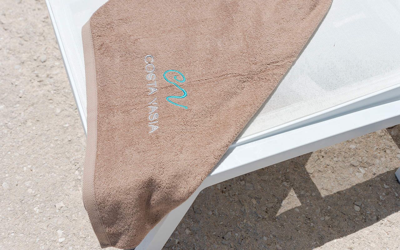 A white lounge chair with a brown towel draped over it, and the text "COSTA YASMIN" visible on the towel.
