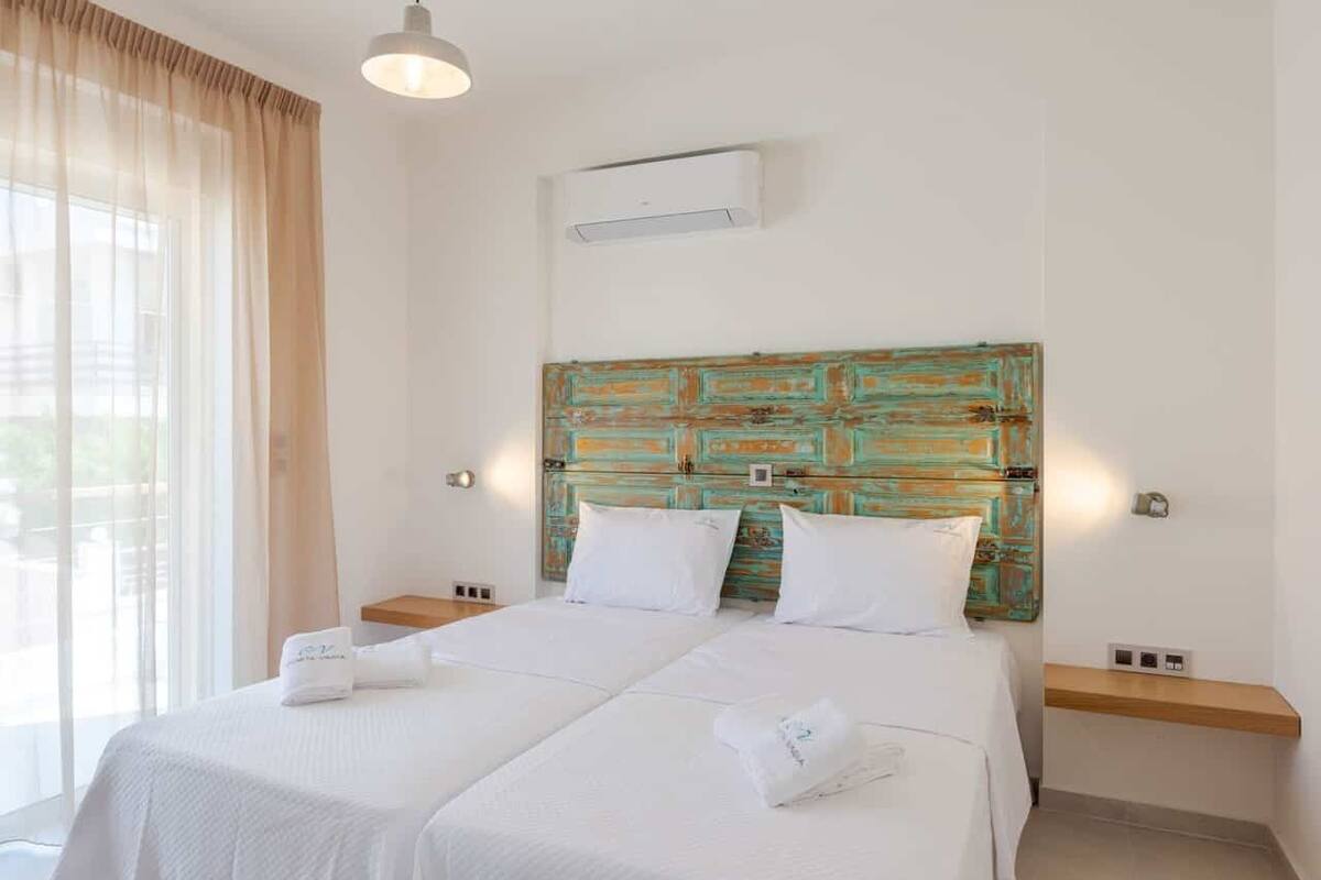Una cama doble con sábanas blancas, un cabecero de madera con un diseño desgastado en tonos aguamarina y naranja, y una mesita de noche con una lámpara.