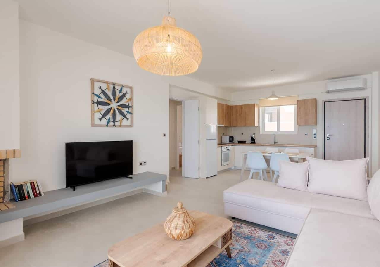 A modern living room with a flat-screen TV, a woven pendant light, a white sofa, and a kitchen in the background.