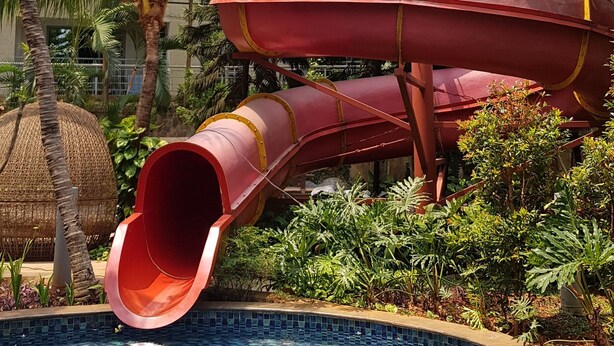 A swimming pool with a slide, surrounded by lush greenery and a multi-story building in the background. - Apatel The Mansion Jasmine Aurora 31 I (Jakarta)