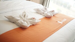 Una cama bien hecha con una colcha blanca y naranja, dos toallas dispuestas en forma de lazo, y una pequeña mesita de noche al lado de la cama.