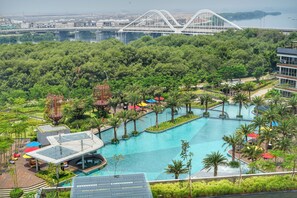 Un resort con una gran piscina rodeada de palmeras y vegetación, un puente moderno al fondo y vistas de un paisaje urbano.