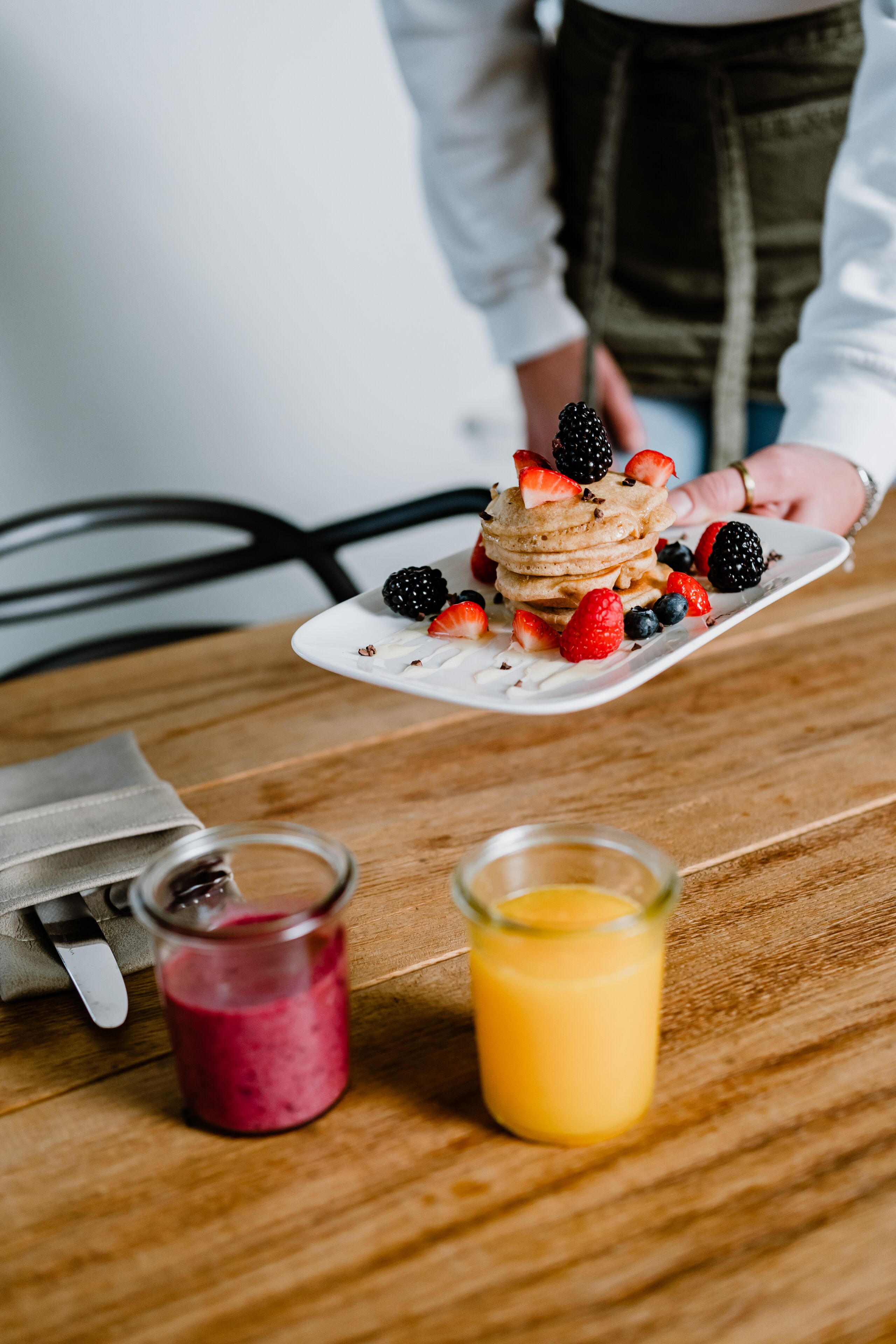 Een ontbijtje met pannenkoeken, belegd met bessen, een glas sinaasappelsap en een glas rode smoothie op een houten tafel.