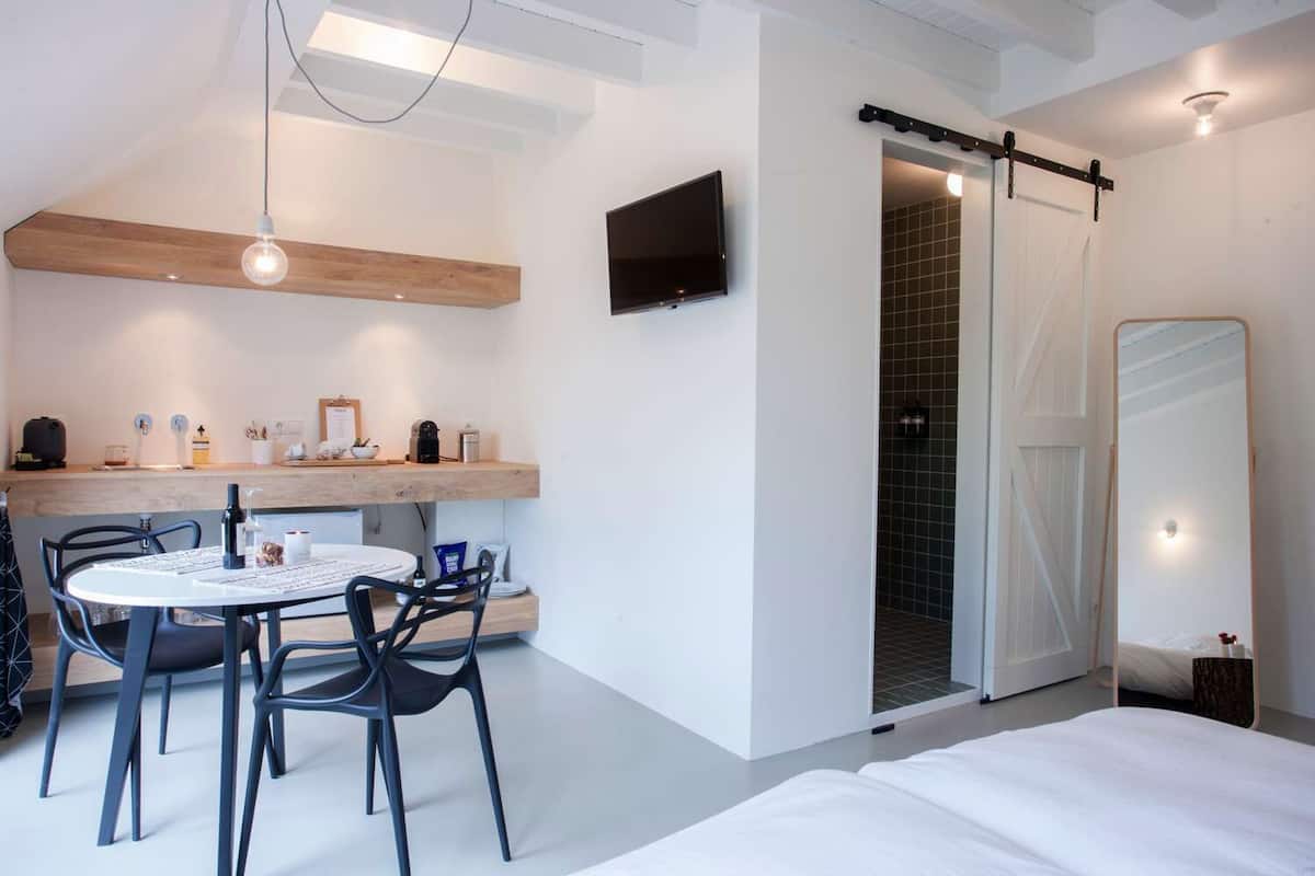 A modern hotel room with a dining area, a TV mounted on the wall, a sliding door leading to a shower, and a bed with white bedding.