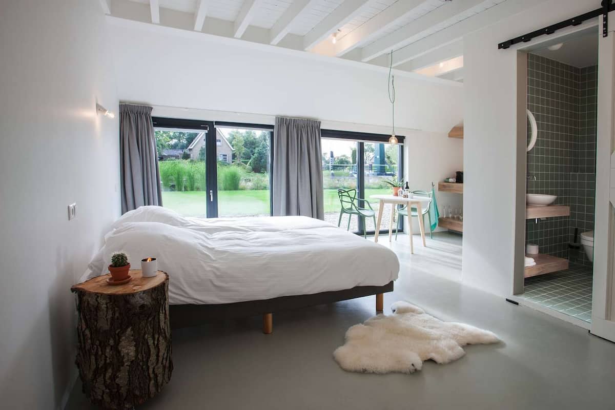 A modern bedroom with a large bed, a wooden side table, a white furry rug, and a view of a garden through large windows.