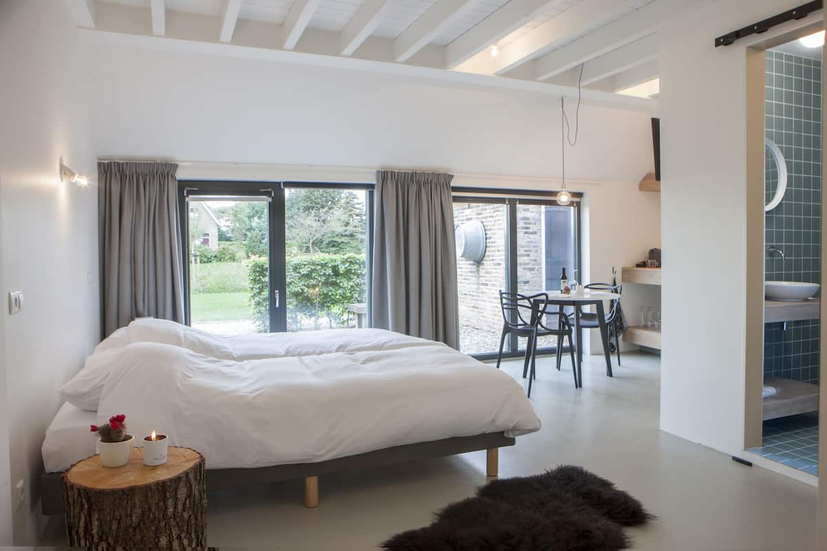 A bedroom with a large bed, a dining table with chairs, and a view of the garden through the window.
