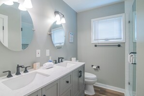 A bathroom with a double sink, a toilet, a large mirror, and a window with blinds.