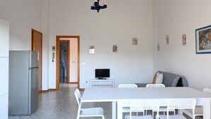 Una cocina moderna con refrigerador, un comedor con mesa y sillas, un televisor sobre un mueble bajo y un pasillo que conduce a otra habitación.