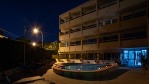Ett hotell med flera våningar, en swimmingpool och en omgivning bestående av solstolar och krukväxter.