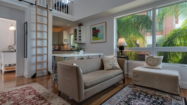 A spacious living room with a high ceiling, a loft bedroom with a wooden ladder, a kitchen with white cabinets, and large windows offering a view of greenery. - Lido Beach Walk! One block to beach! (Sarasota)