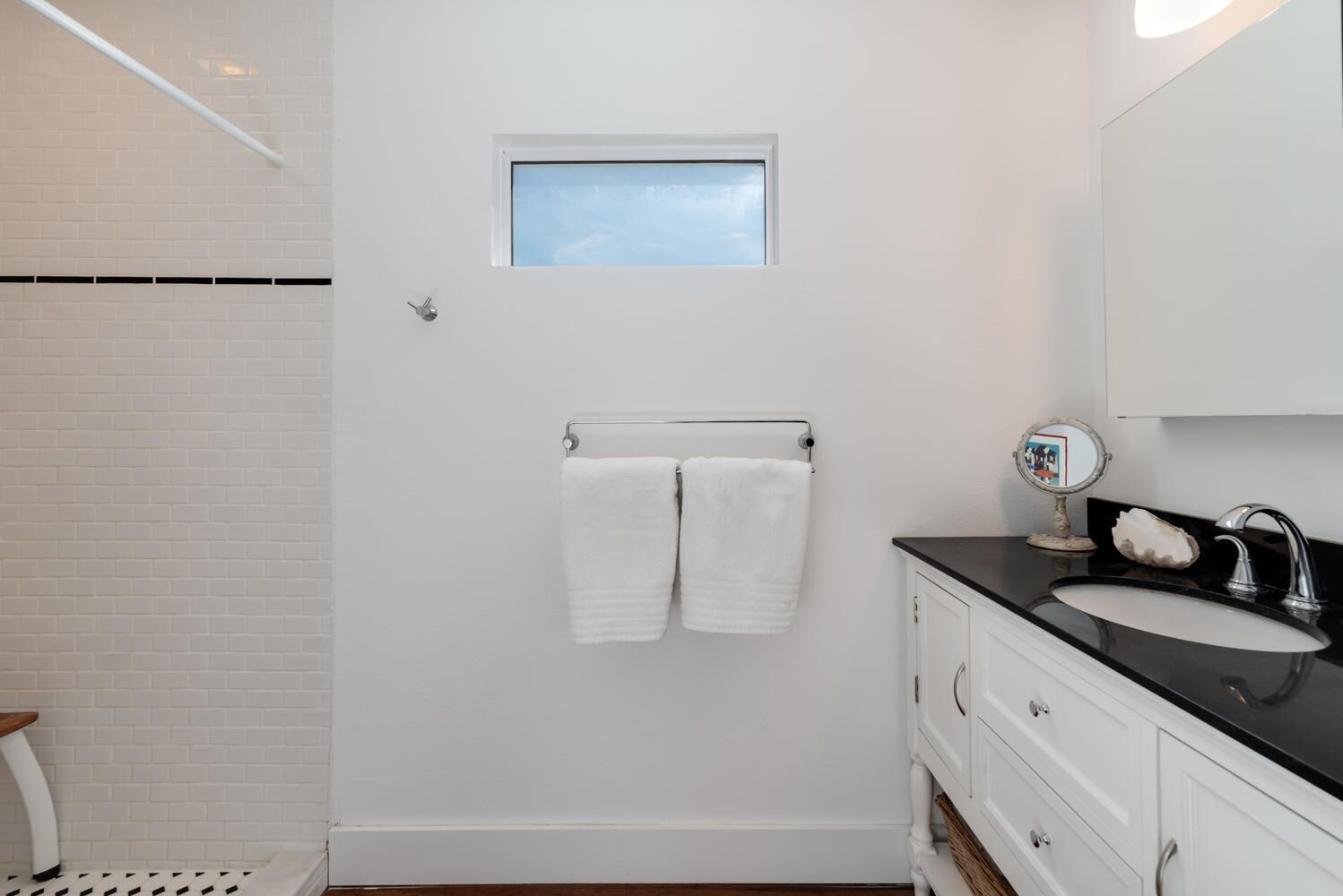 Une salle de bain avec un mur recouvert de carreaux blancs, une petite fenêtre, un porte-serviettes avec deux serviettes, un lavabo avec un miroir et un meuble.