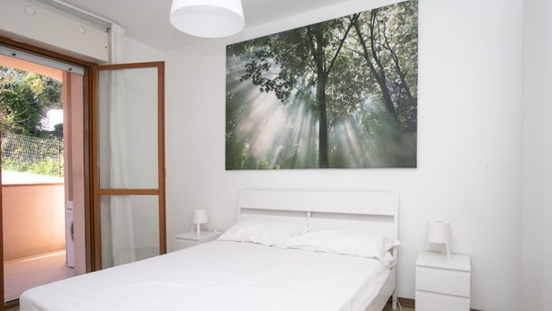 A bedroom with a bed, two bedside tables, a nightstand, a window with curtains, and a painting of a forest.