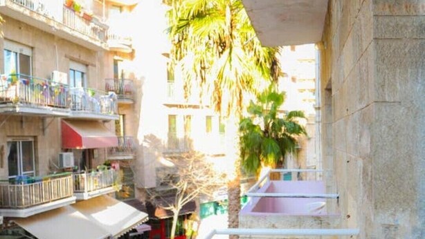 A balcony with a wicker chair, a view of a street with buildings and a palm tree, and a concrete wall. - Great Studio in amazing location (Jérusalem)