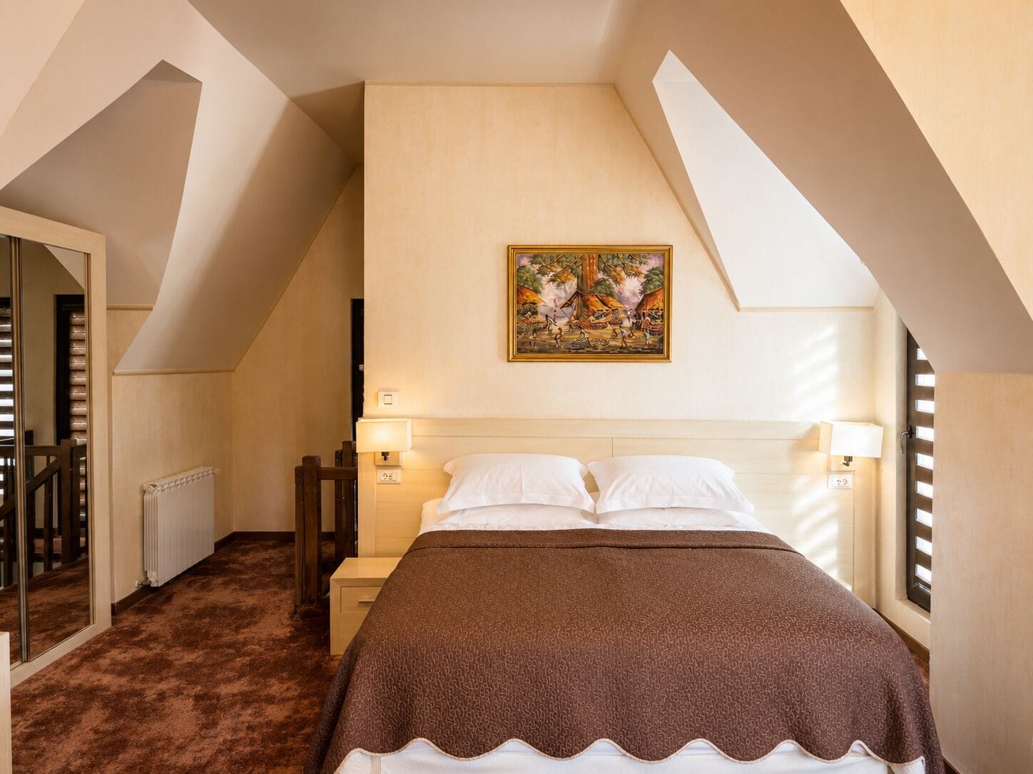 A bedroom with a bed, two bedside lamps, a radiator, a wooden nightstand, a painting on the wall, and a sloped ceiling.