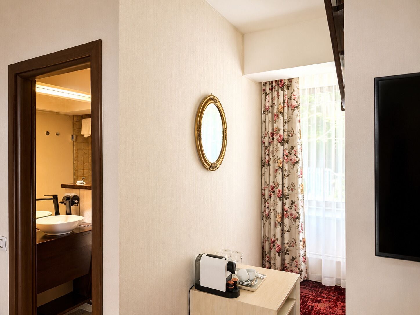 A hotel room with a bathroom, a mirror, a small table with a coffee maker, and a window with floral curtains.