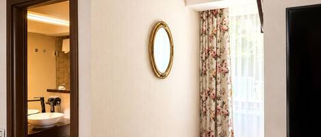 A hotel room with a bathroom, a mirror, a small table with a coffee maker, and a window with floral curtains.