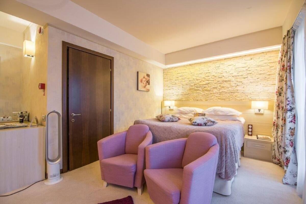 Ein Schlafzimmer mit einem Bett, zwei Sesseln, einem Nachttisch, einer Lampe, einem Wandbild und einer Tür.
