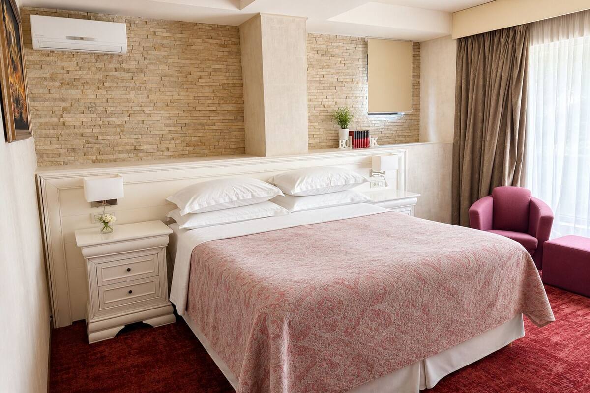 Ein Hotelzimmer mit Backsteinwand, einem Bett mit gemustertem Überwurf, einem Nachttisch mit Lampe, einem Sessel und einem Fenster mit Vorhängen.