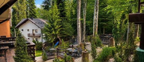 A rustic outdoor dining area with wooden tables and chairs, surrounded by trees and a small building with a balcony.