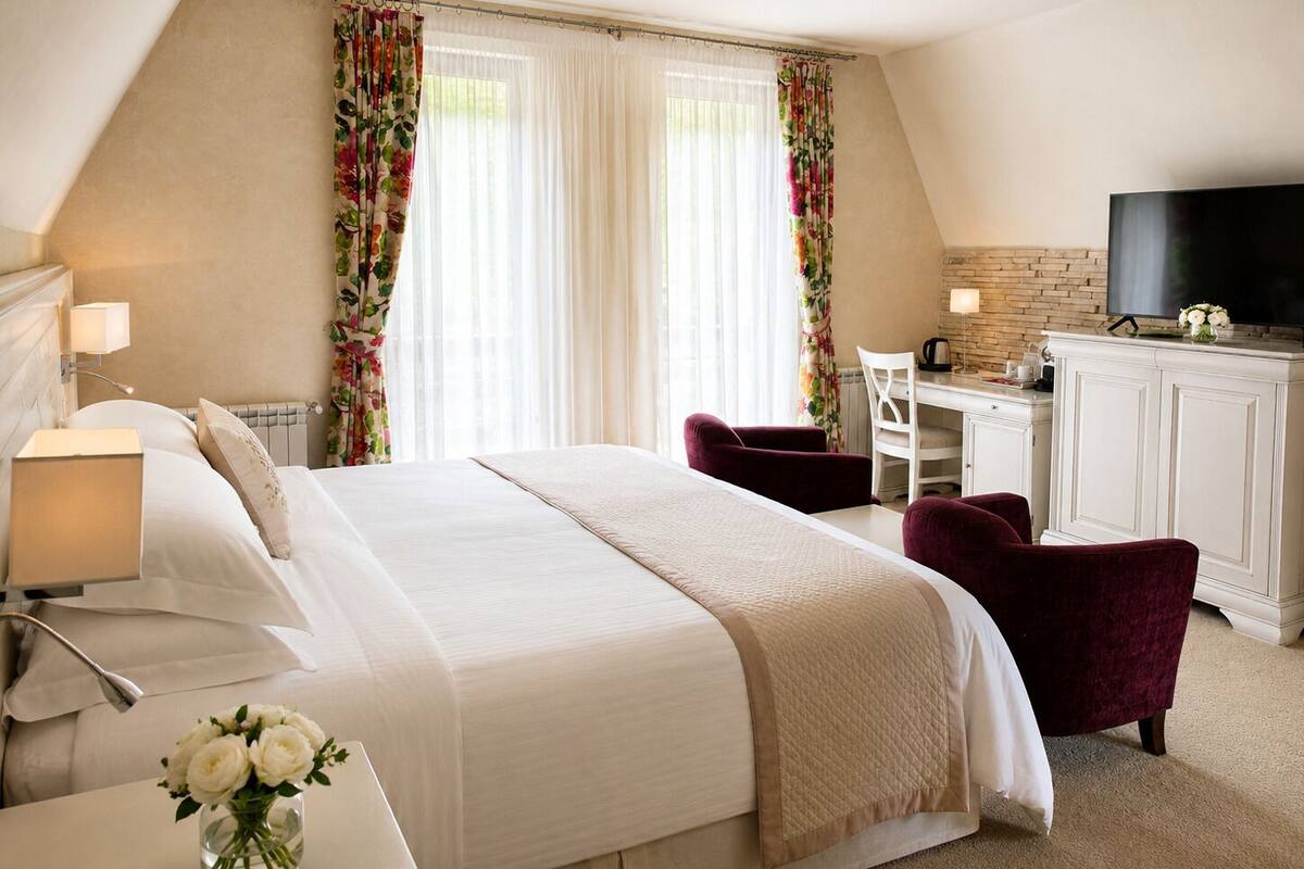 Ein Hotelzimmer mit Bett, Schreibtisch, Stuhl, Fernseher und einer Vase mit Blumen.
