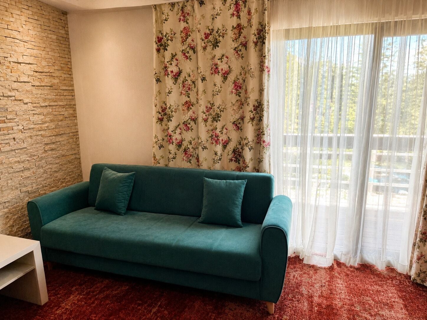 A teal sofa with two cushions, a white side table, a brick-patterned wall, floral curtains, and a window with sheer white curtains.