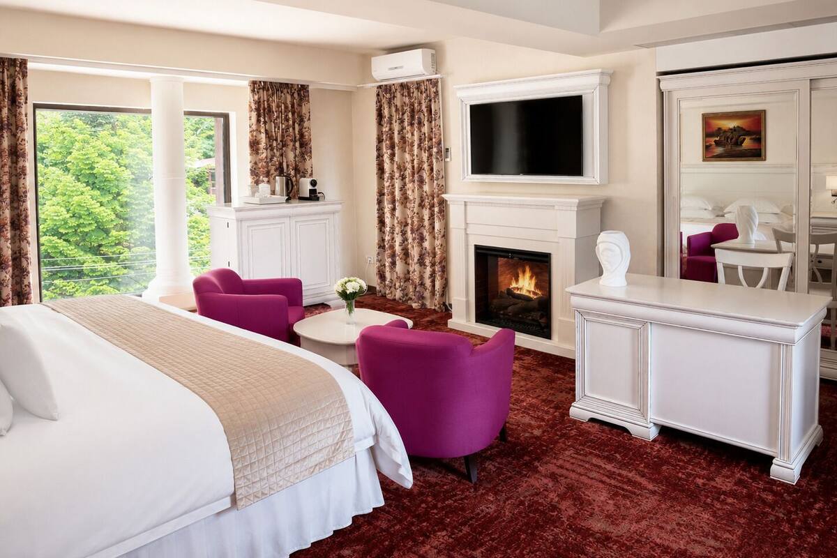 Ein Hotelzimmer mit Bett, Kamin, Fernseher, Schreibtisch und Blick auf Grün.