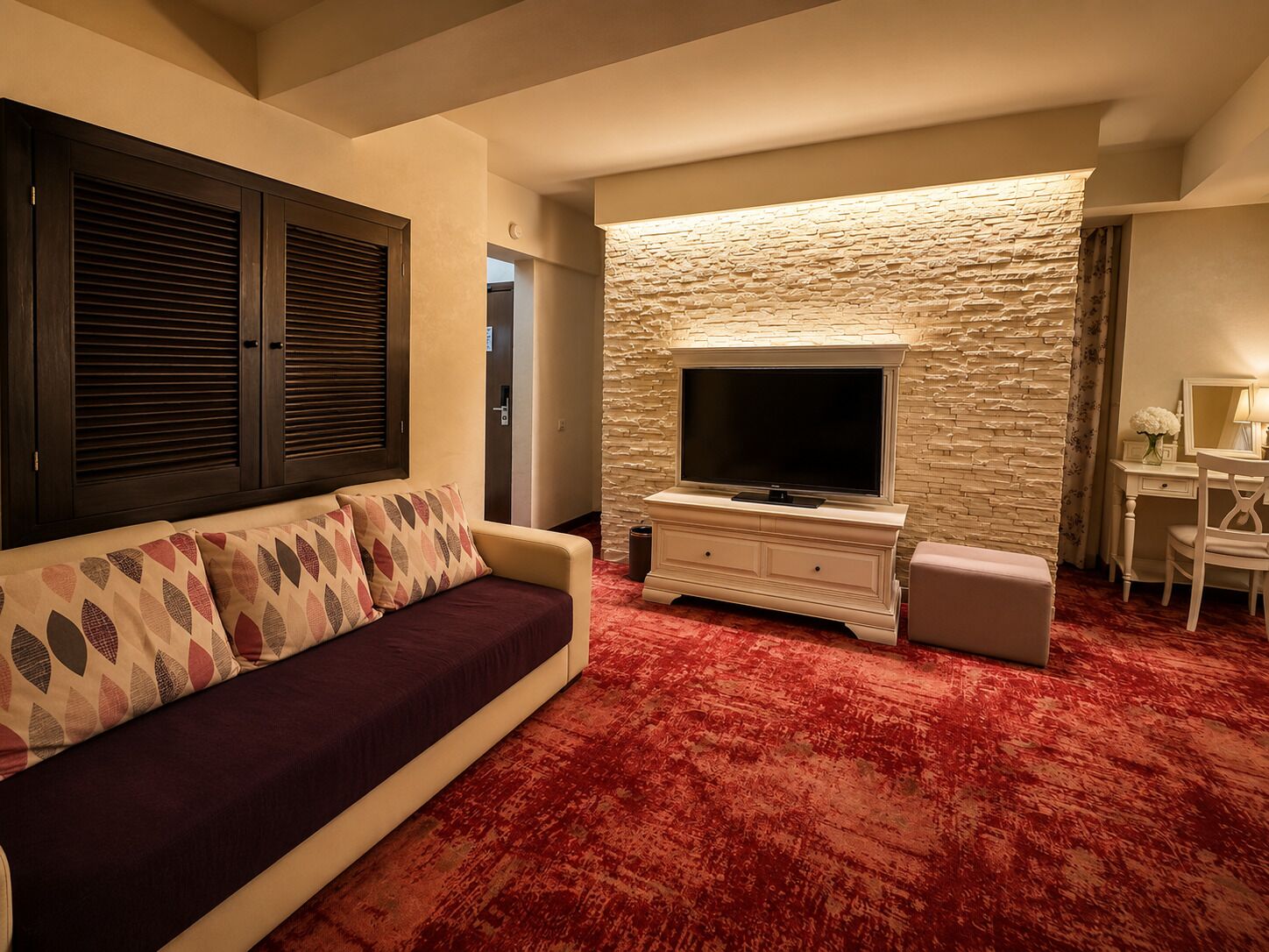 A room with a sofa, a TV on a white cabinet, a stone wall, and a desk with a lamp.