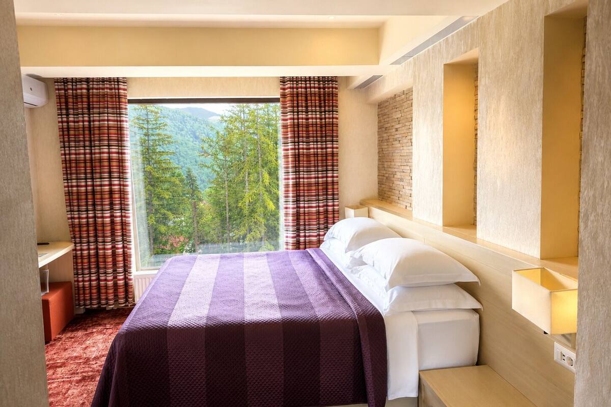Ein Schlafzimmer mit einem Bett, einem Fenster mit Vorhängen, einem Nachttisch und einer Wandlampe.