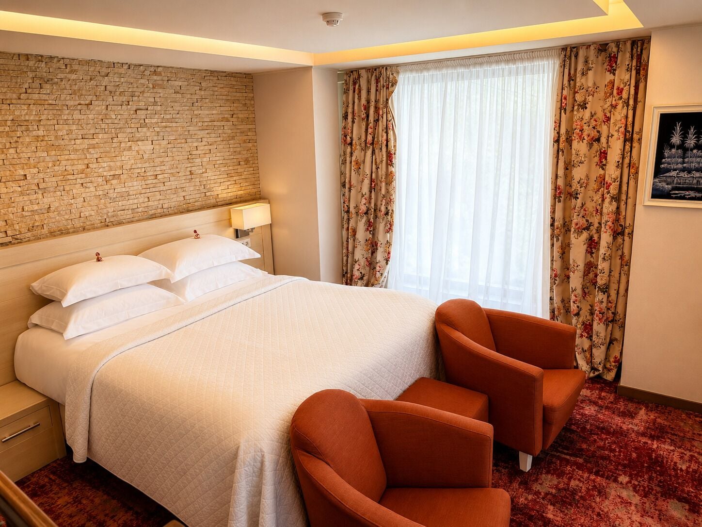 A hotel room with a brick wall, a large bed with white bedding, two orange armchairs, a window with floral curtains, and a framed picture on the wall.