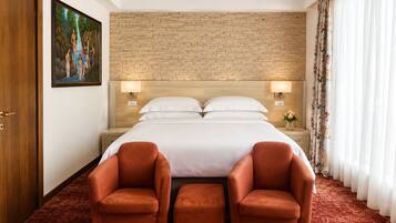 Habitación de hotel con pared de ladrillo, cama con ropa blanca, dos sillones rojos y un cuadro en la pared.