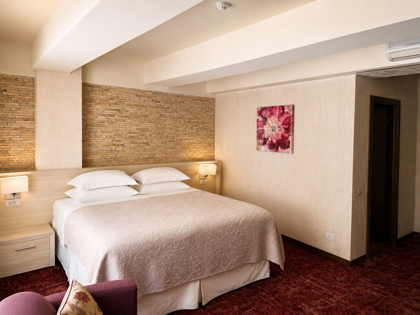 Ein Hotelzimmer mit Bett, Nachttischlampen, einer Wand aus Ziegelsteinen, einem Blumenbild und einer Tür.