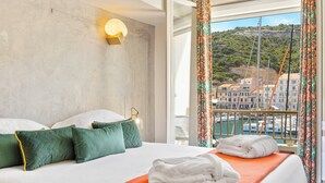 A hotel room with a bed, pillows, a nightstand, a window with curtains, and a view of a harbor. - Hotel Solemare (Bonifacio)