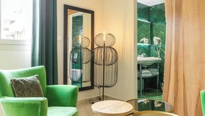 A hotel room with a green armchair, a floor lamp, a round table, and a bathroom with a green herringbone tile backsplash. - Hotel Solemare (Bonifacio)