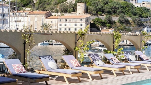 A pool area with lounge chairs, a clear view of a historic stone fortress in the background, and a bridge connecting the pool to the shore. - Hotel Solemare (Bonifacio)