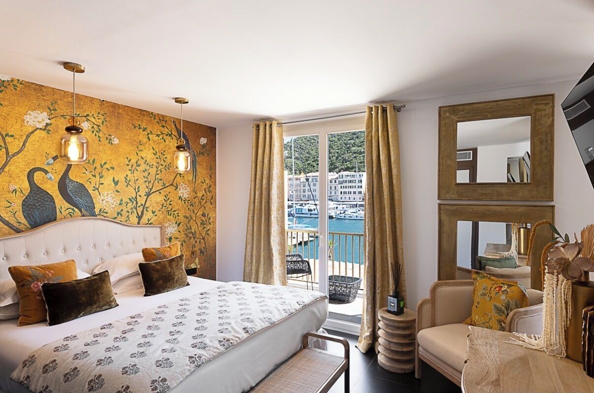 A bedroom with a bed, a large window with curtains, a chair, a mirror, and a wall with a peacock painting.