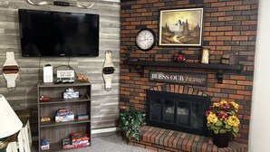 A living room with a brick fireplace, a flat-screen TV mounted on the wall, and a bookshelf filled with board games.