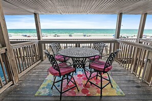Eine Holzterrasse mit rundem Tisch und Stühlen mit Blick auf einen Strand mit Sonnenschirmen und Menschen.