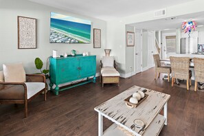 A living room with a teal cabinet, wicker chairs, a white table, and a large painting of a beach.