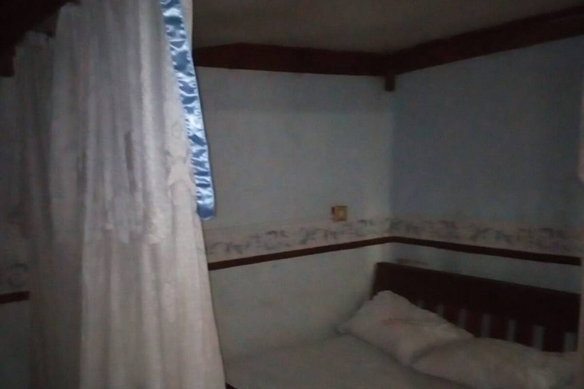 Una habitación tenuemente iluminada con una cama individual, cabecera de madera y cortina entreabierta.