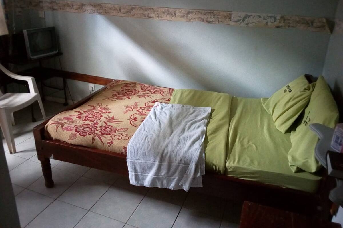 Una cama individual con ropa de cama de estampado floral, una toalla blanca y almohadas verdes en una habitación con piso de baldosas y un televisor en una esquina.