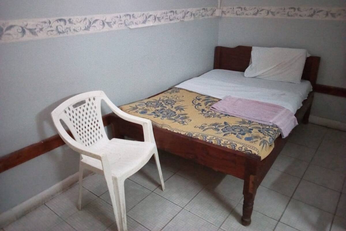 Una habitación pequeña con cama individual, una cama de madera, una silla de plástico blanco y una colcha estampada.