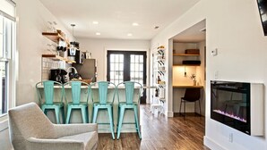 Un salon moderne avec une cheminée, un coin repas avec des chaises de couleur turquoise et une cuisine avec des meubles blancs et un réfrigérateur noir.