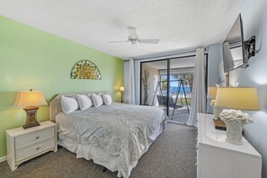 Une chambre à coucher comprenant un lit, des tables de chevet, un ventilateur de plafond, une grande fenêtre avec des rideaux et une vue sur l’extérieur.