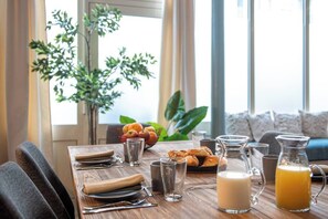 A breakfast table set with croissants, milk, juice, and glasses, with a plant and a bowl of fruit in the background. - My Cottage in Paris (Paris)