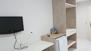 A modern, minimalist room with a flat-screen TV mounted on the wall, a small refrigerator, and a white desk with a chair. - Hotel Portinari (Vilhena)