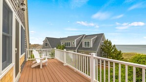A wooden deck with white railings and two Adirondack chairs, overlooking a body of water and a row of houses. - Gorgeous home sleeping 12 with ocean views (West Harwich)
