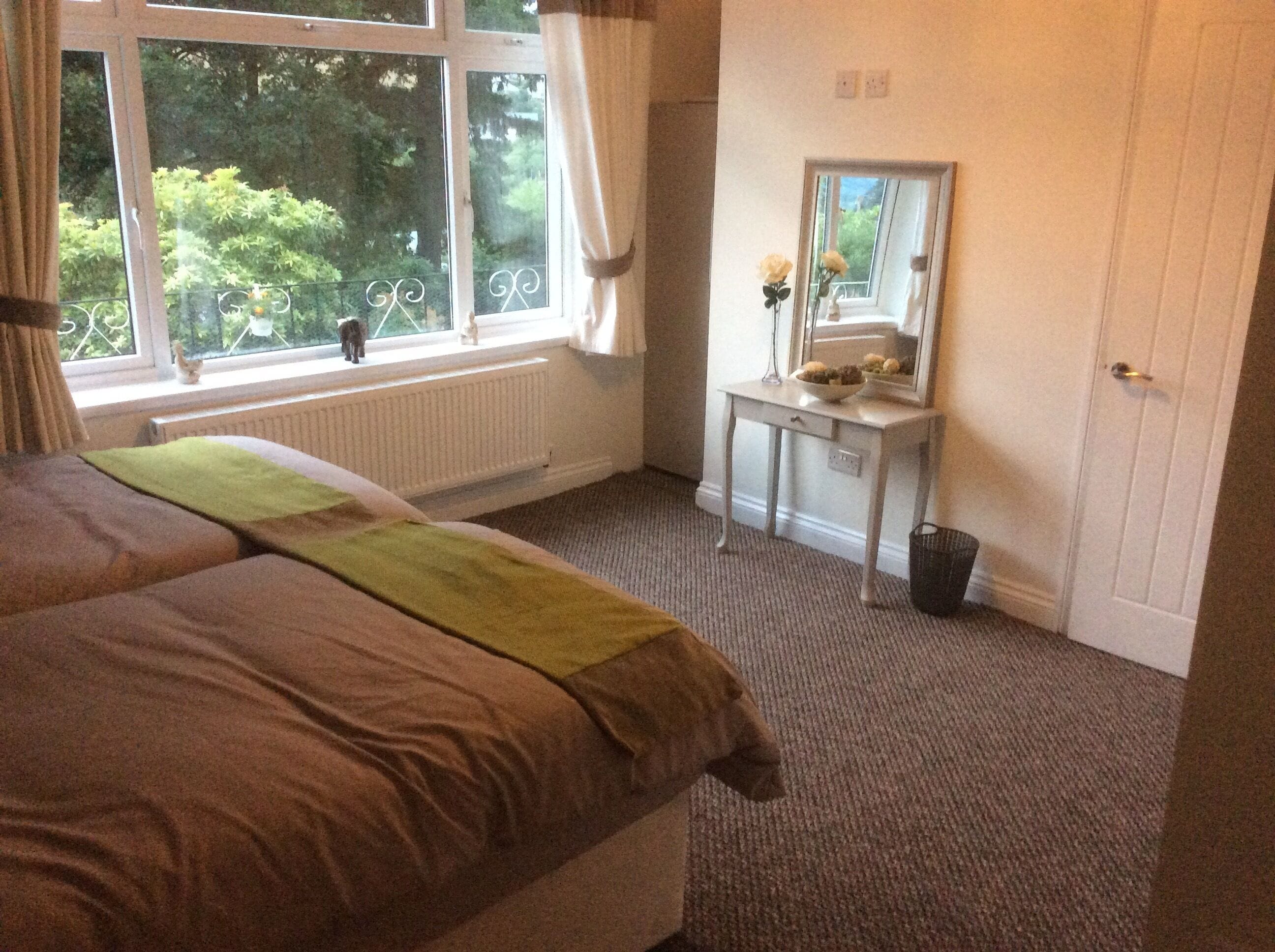 A bedroom with a bed, a window with a view of greenery, a bedside table with a mirror, and a radiator.