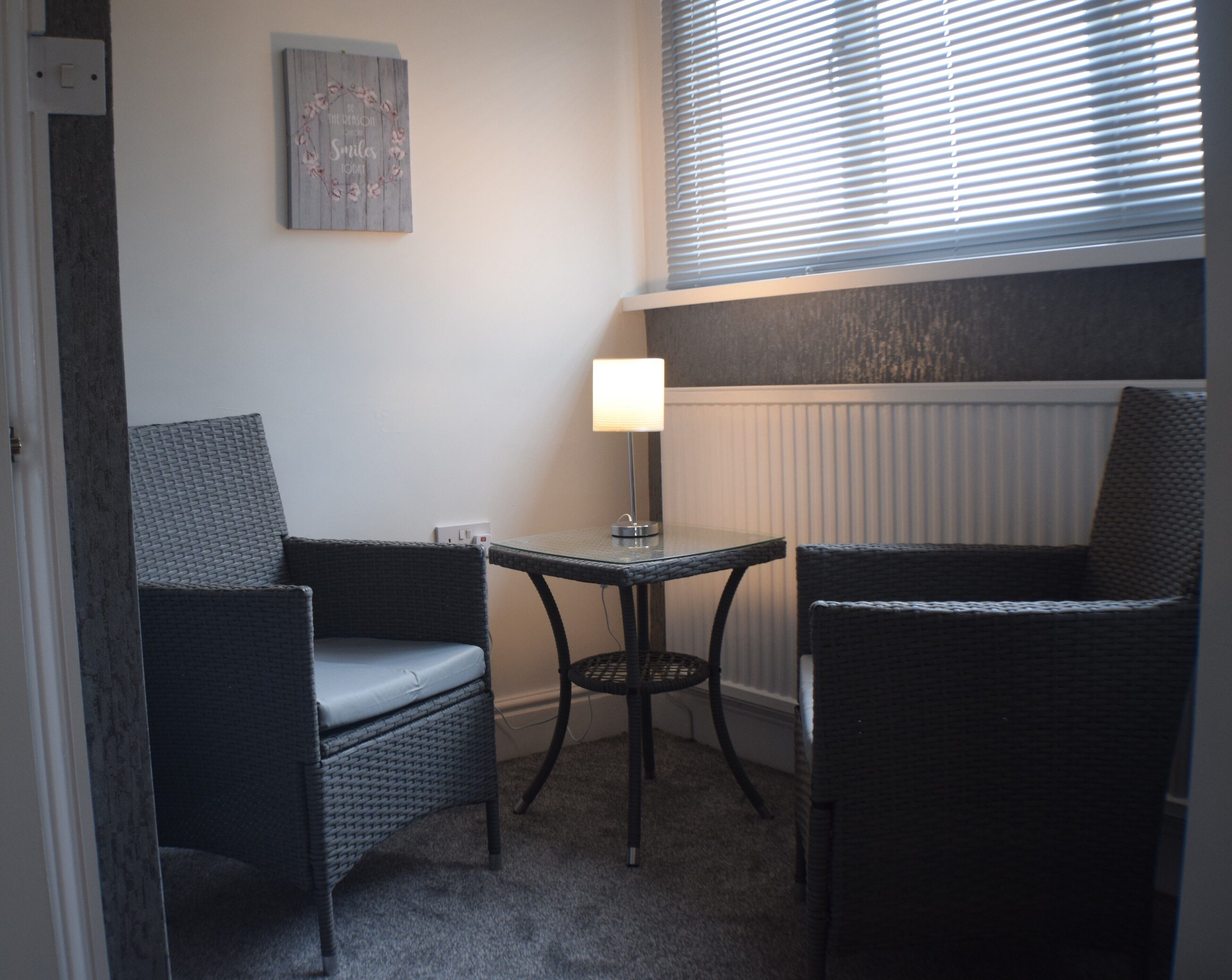 Et lille rum med to fletstole, et bord, en lampe og en radiator.